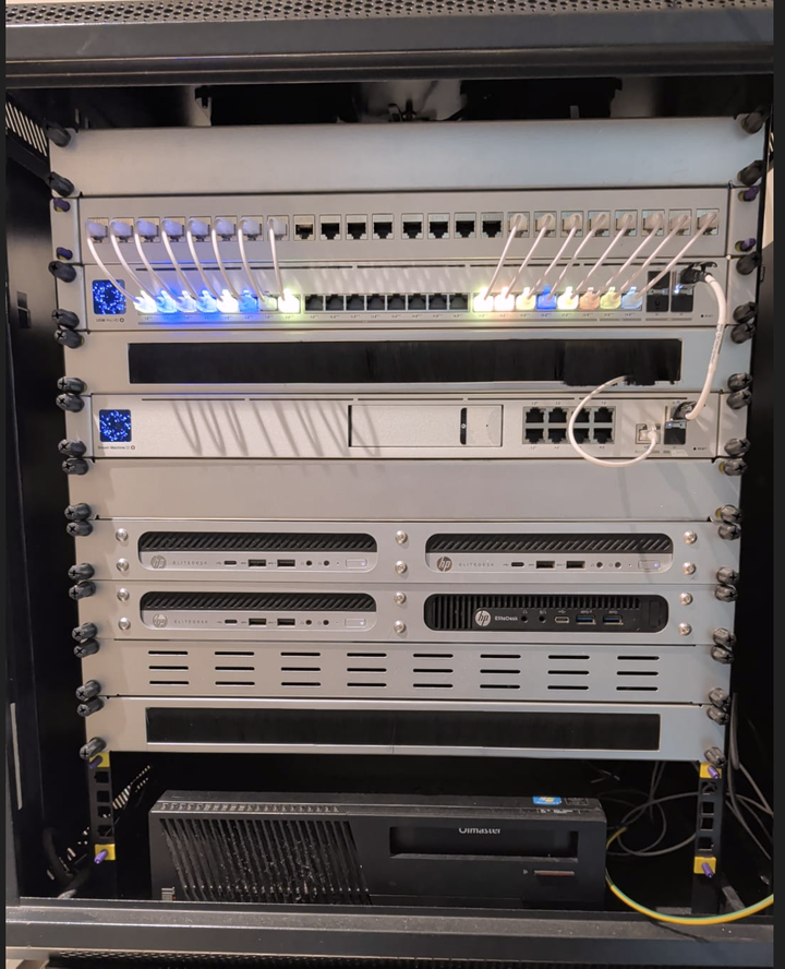 My 12U homelab rack: patch panel and UniFi switch with lit ports at the top, a UniFi Dream Machine SE below, and HP EliteDesk mini PCs on shelves beneath.